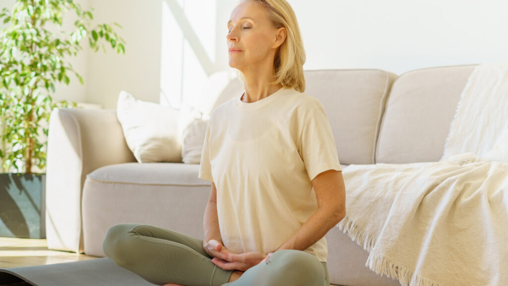 אישה מבוגרת יושבת בסלון על הרצפה עושה מדיטציה